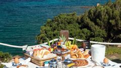 Frühstückstisch auf einer Terrasse mit traumhaftem Blick auf das tiefblaue Meer – serviert werden frische Croissants, Brötchen, Obst, Säfte und Kaffee – ein perfekter Start in den Tag mit mediterranem Panorama.