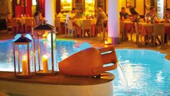 Lipari Hotel Arciduca Restaurant ©michelangelo int travel, momento Stimmungsvoll beleuchteter Poolbereich mit Wasserspiel in Tonkrugform und dekorativen Laternen, im Hintergrund ein elegantes Restaurant mit Gästen bei Abendessen.