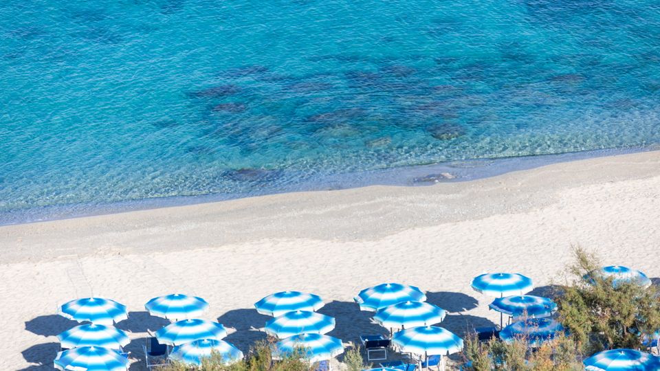 Reihe von blau-weißen Sonnenschirmen an einem feinsandigen Strand mit kristallklarem Wasser in verschiedenen Türkistönen.