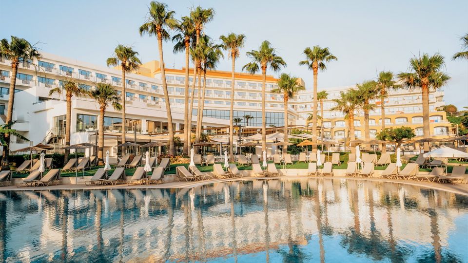 Großzügiger Poolbereich mit Liegen unter Palmen – ideal zum Entspannen und Genießen der Sonne direkt vor dem eleganten Hotelgebäude.
