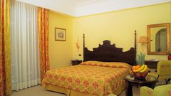 Lipari Hotel Arciduca Zimmerbeispiel ©michelangelo int travel, momento Klassisch eingerichtetes Hotelzimmer mit großem Doppelbett, floralen Vorhängen und stilvollen Möbeln in warmen Gelb- und Rottönen – auf dem Tisch eine Obstschale mit frischen Früchten.