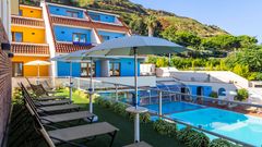 Moderne Hotelanlage mit bunten Fassaden, sonniger Liegewiese und großzügigem Poolbereich inmitten grüner Hügellandschaft.