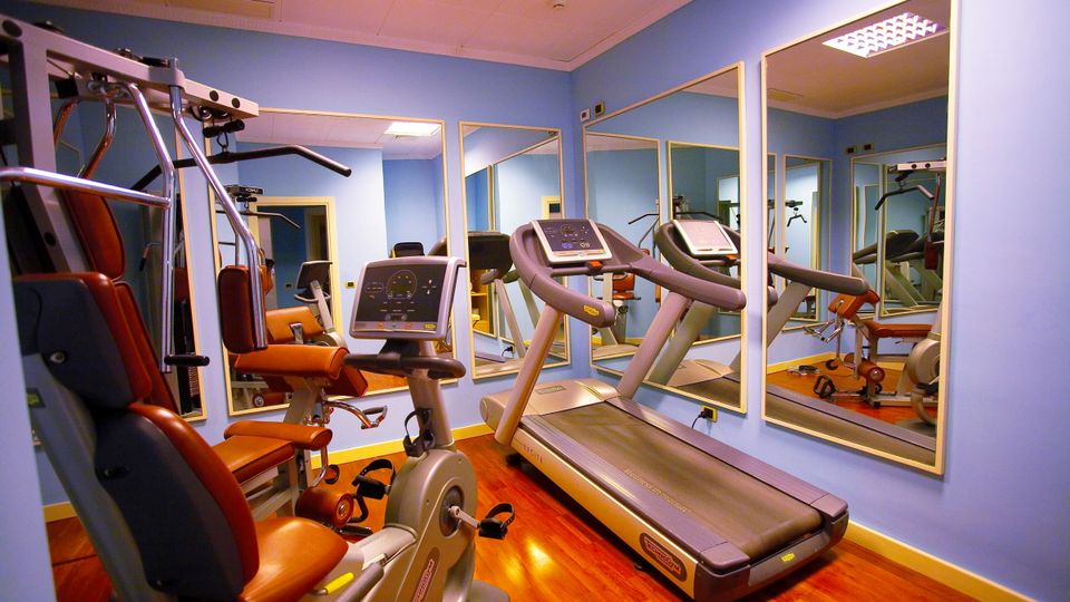 Fitnessbereich Hotel Arciduca ©michelangelo int travel, momento Kompakt ausgestatteter Fitnessraum mit Laufband, Ergometer, Kraftstation und großen Spiegeln – moderne Geräte auf glänzendem Holzfußboden in freundlicher Atmosphäre.